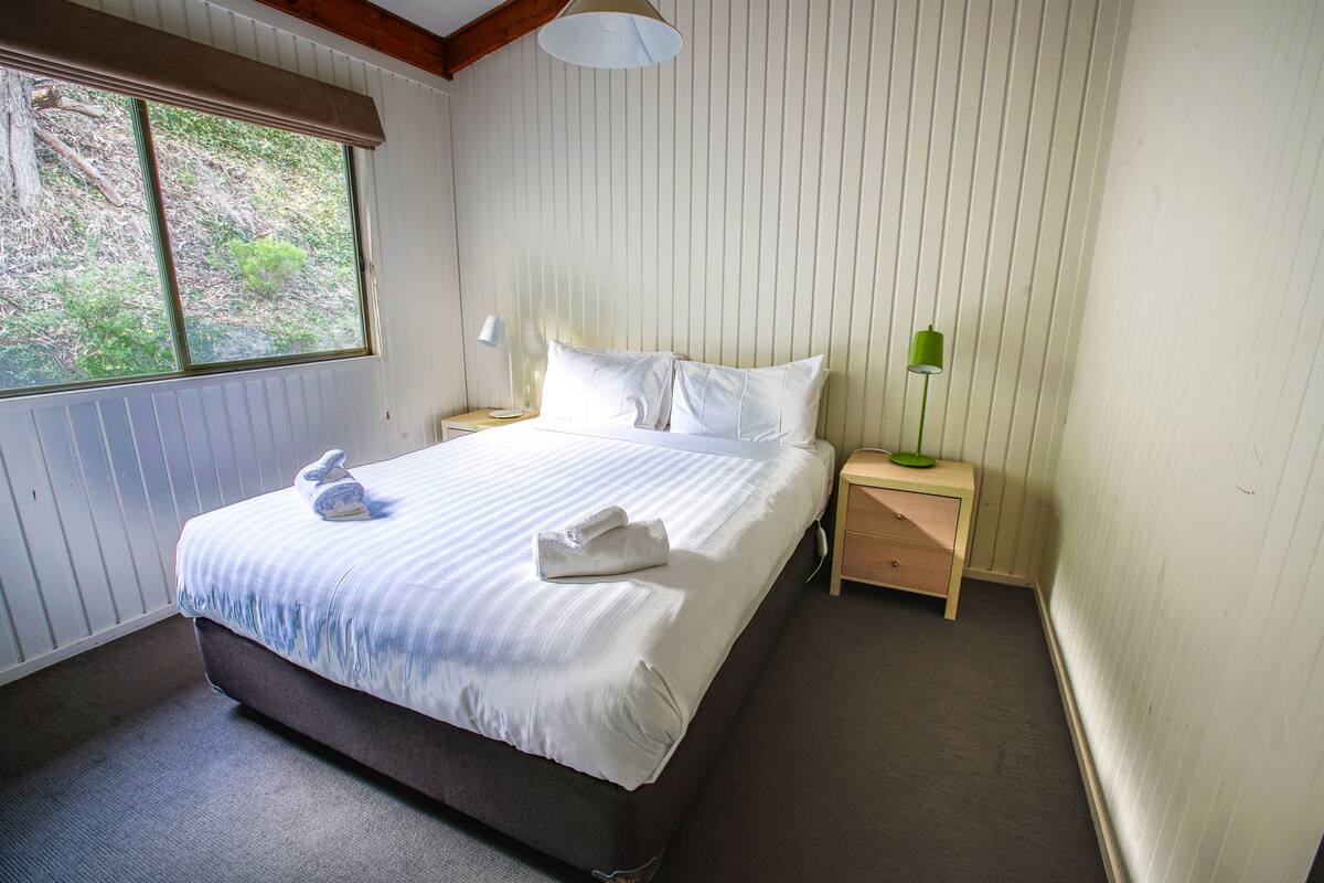 Un lit bien fait, avec du linge de lit blanc et deux serviettes, une lampe de chevet et une table de nuit.