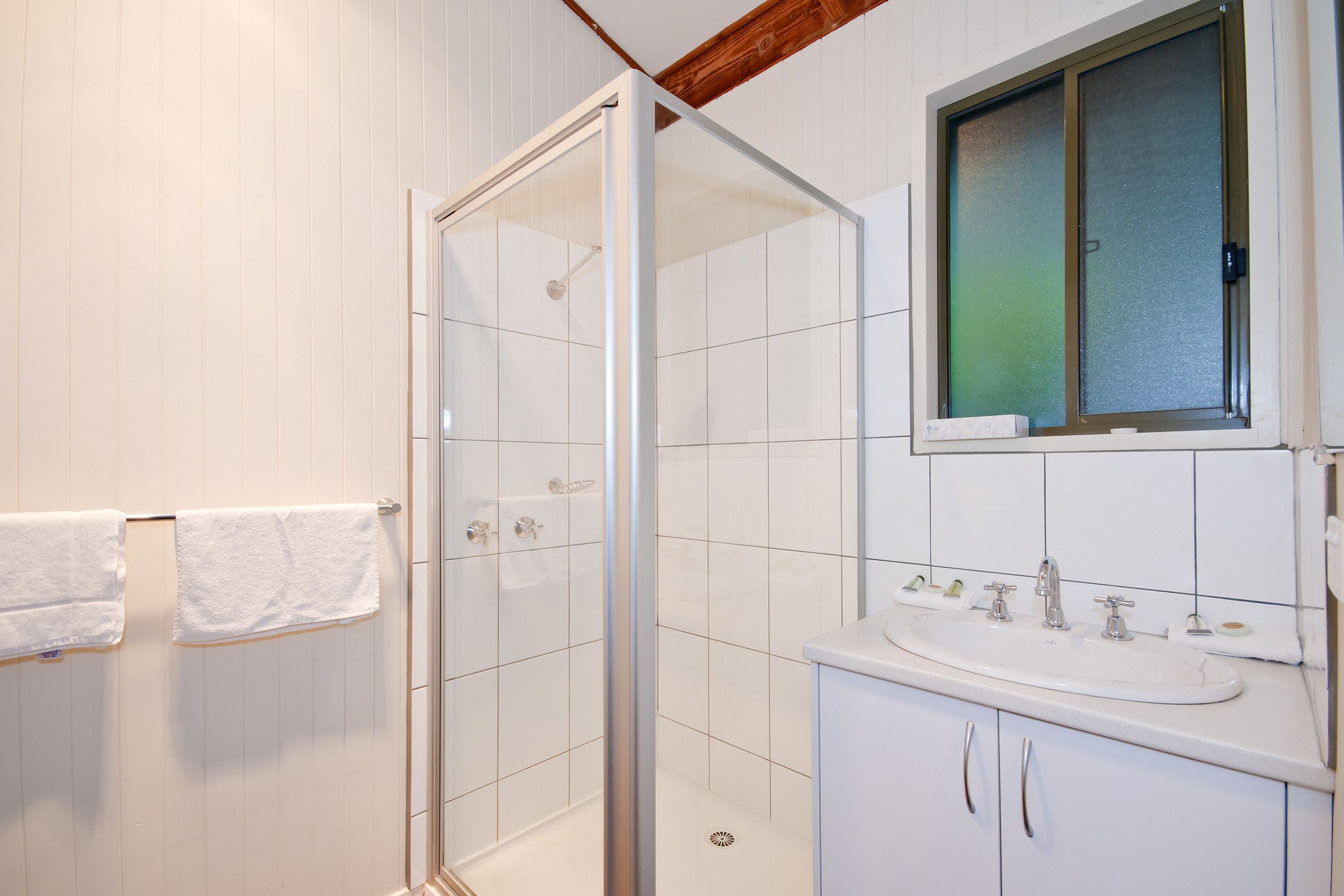 A bathroom with a glass shower enclosure, a white sink, and a window above the mirror.