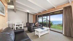 A modern living room with a sofa, a coffee table, and a dining area with chairs. There is a large window offering a view of the sea. - Beacon Point Ocean View Villas (Skenes Creek)