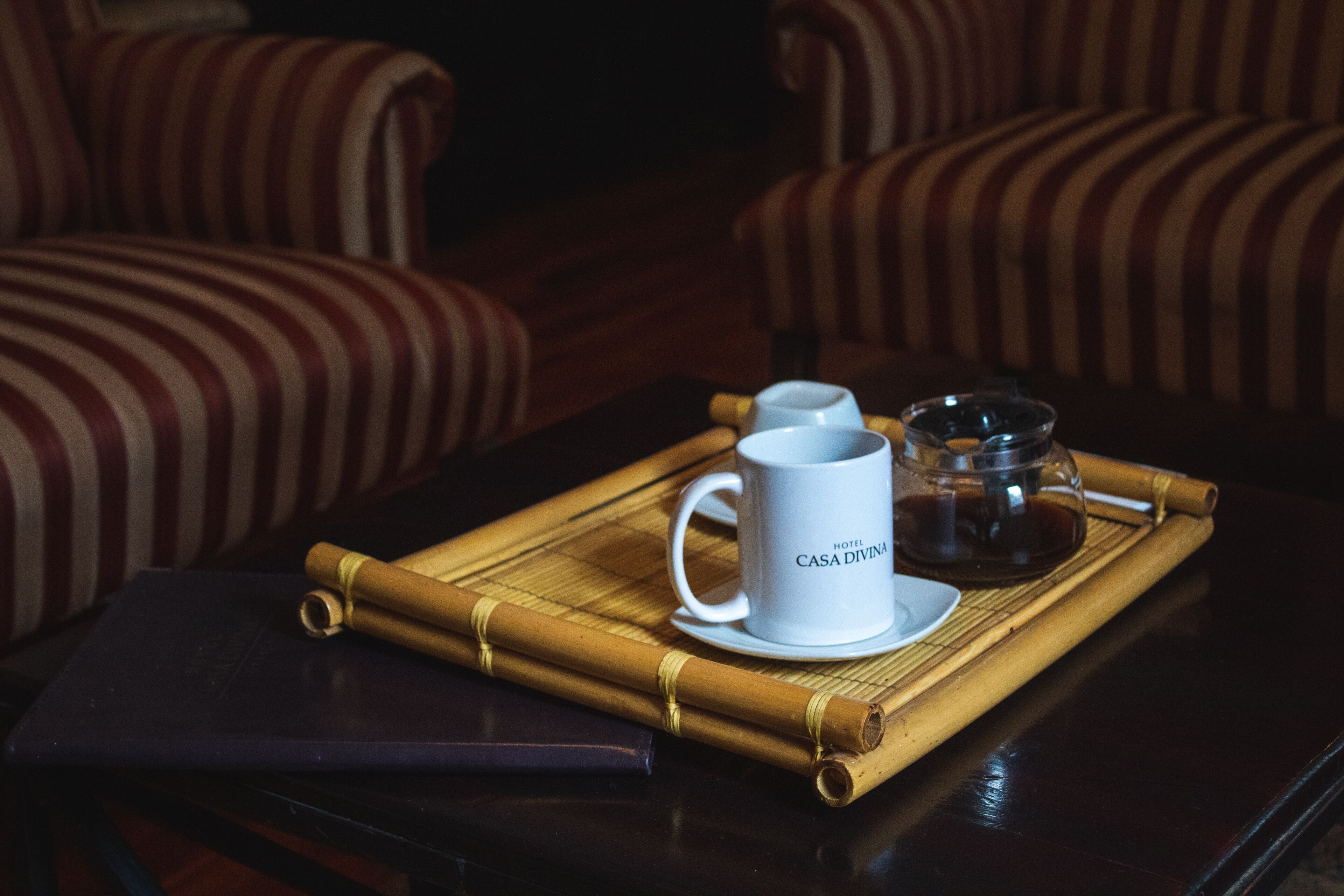 A room with striped upholstered chairs, a dark wooden table, a white mug with "HOTEL CASA DIVINA" text, a glass teapot, and a bamboo mat.