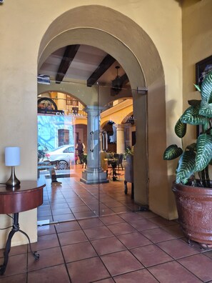 A hotel lobby with a tiled floor, a large potted plant, a lamp, and a glass door leading to another area.