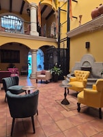 Um hall espaçoso com teto alto, piso de azulejos e diversas opções de assento, incluindo poltronas e sofás. Há plantas em vasos e uma fonte decorativa.