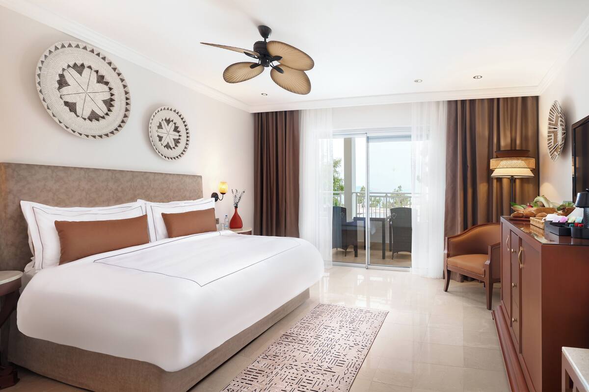 A hotel room with a large bed, a ceiling fan, a desk with a lamp, a chair, and a view of the outdoors.