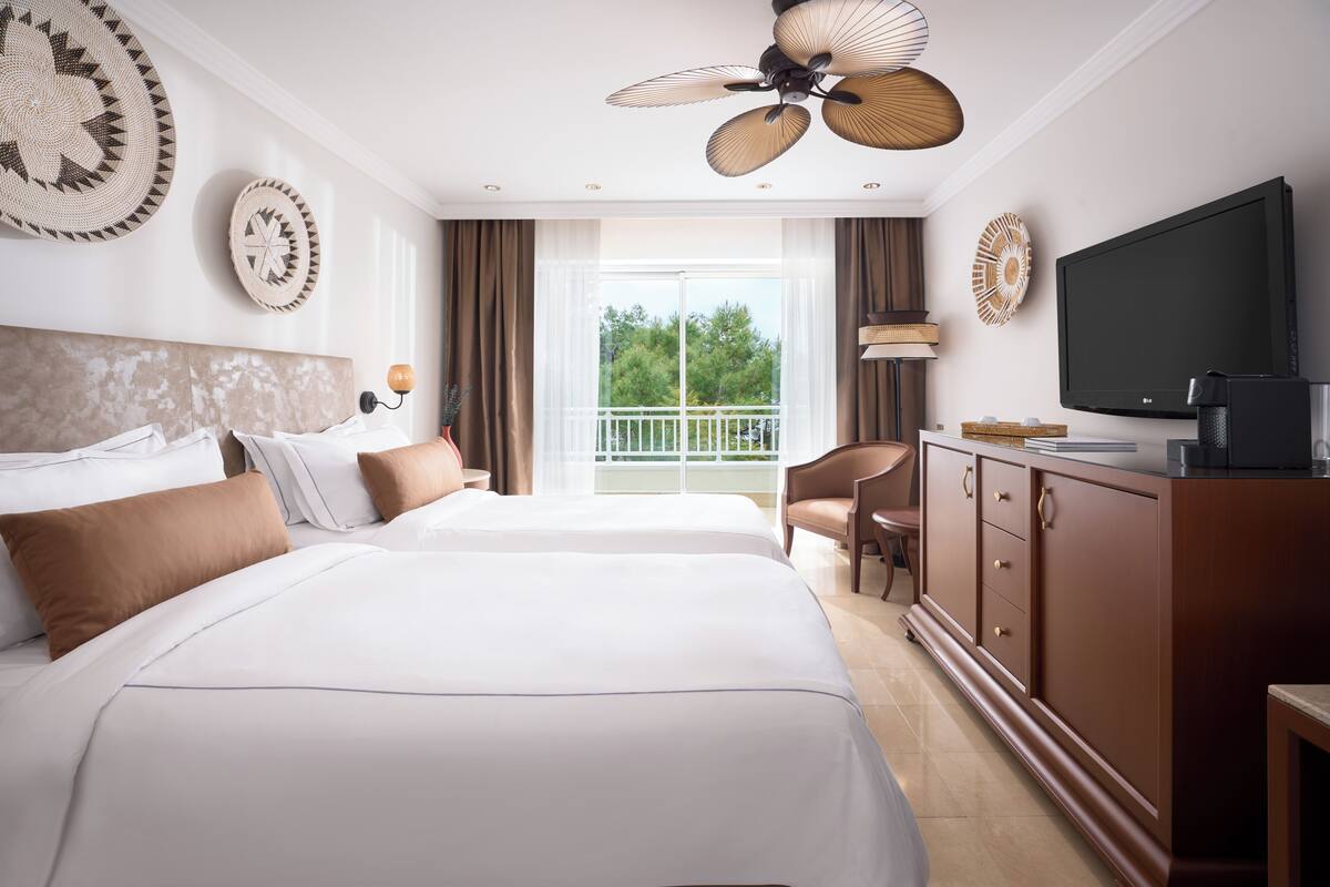 A hotel room with two beds, a TV, a ceiling fan, a balcony with a view, and a wooden cabinet.