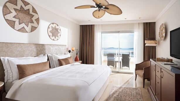A hotel room with a large bed, a ceiling fan, a TV, a balcony with a view, and a dining area.