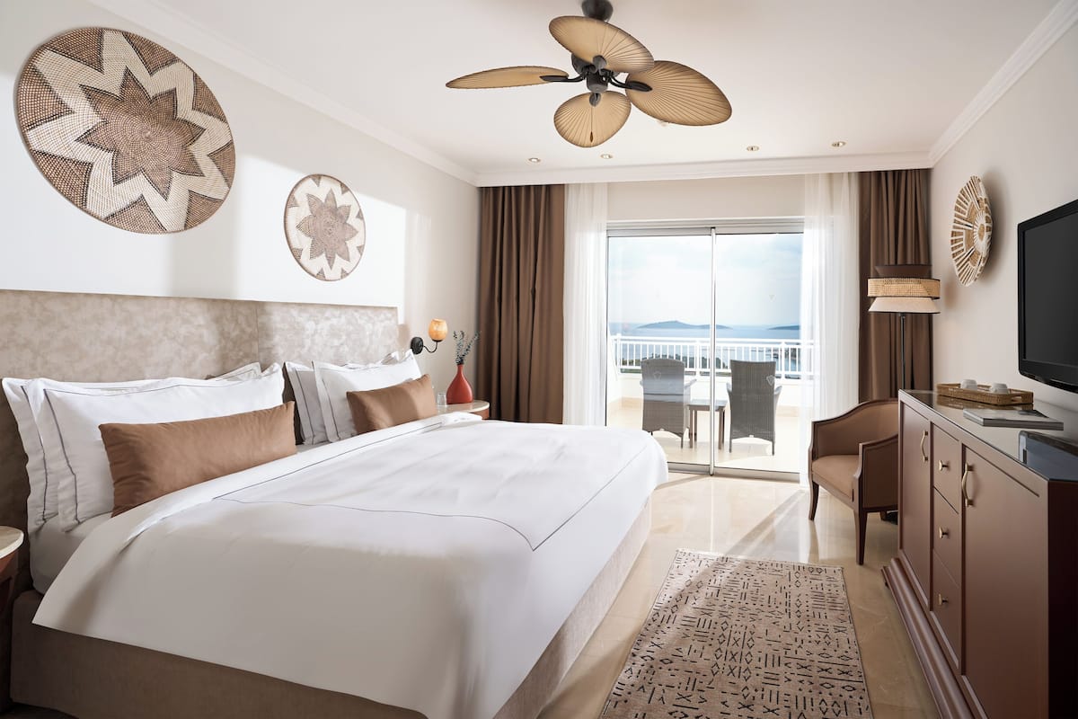 A hotel room with a large bed, a ceiling fan, a TV, a balcony with a view, and a dining area.