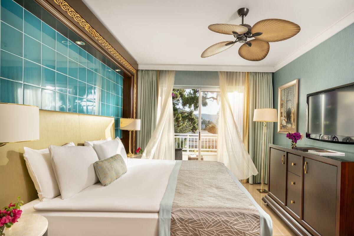 A hotel room with a large bed, a ceiling fan, a TV, and a balcony with curtains.