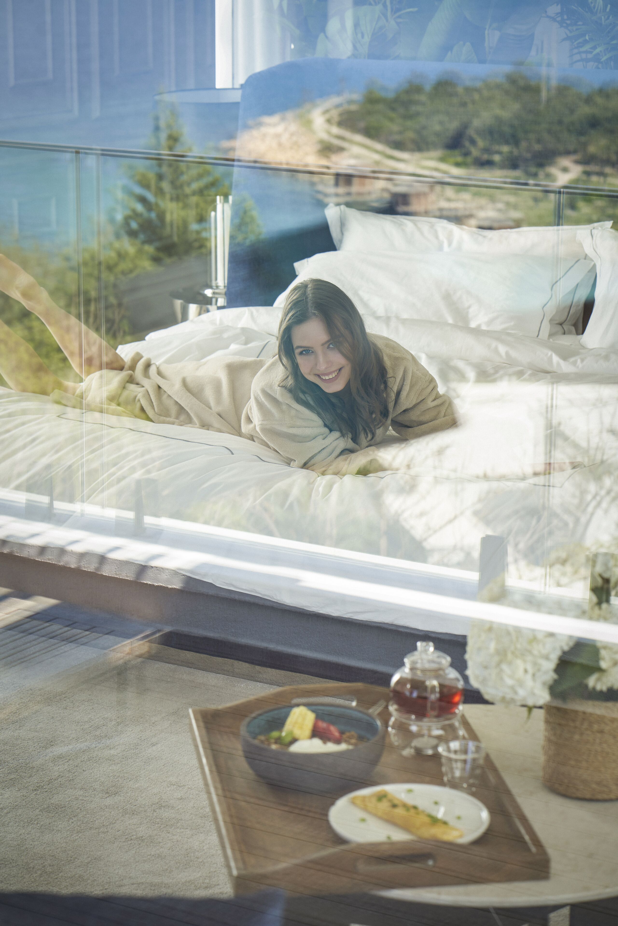 A bed with a view of a landscape through a window, a bedside table with a bowl of fruit, a glass of water, and a teapot.