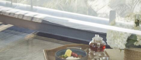 A bed with a view of a landscape through a window, a bedside table with a bowl of fruit, a glass of water, and a teapot.