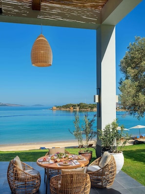 Un área de comedor al aire libre techada, con mobiliario de mimbre, una mesa preparada para una comida y vistas al mar y la costa lejana.