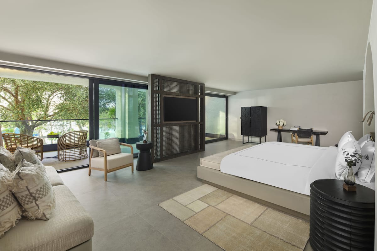 A modern bedroom with a large bed, a sofa, a dining table, and a view of greenery through large windows.