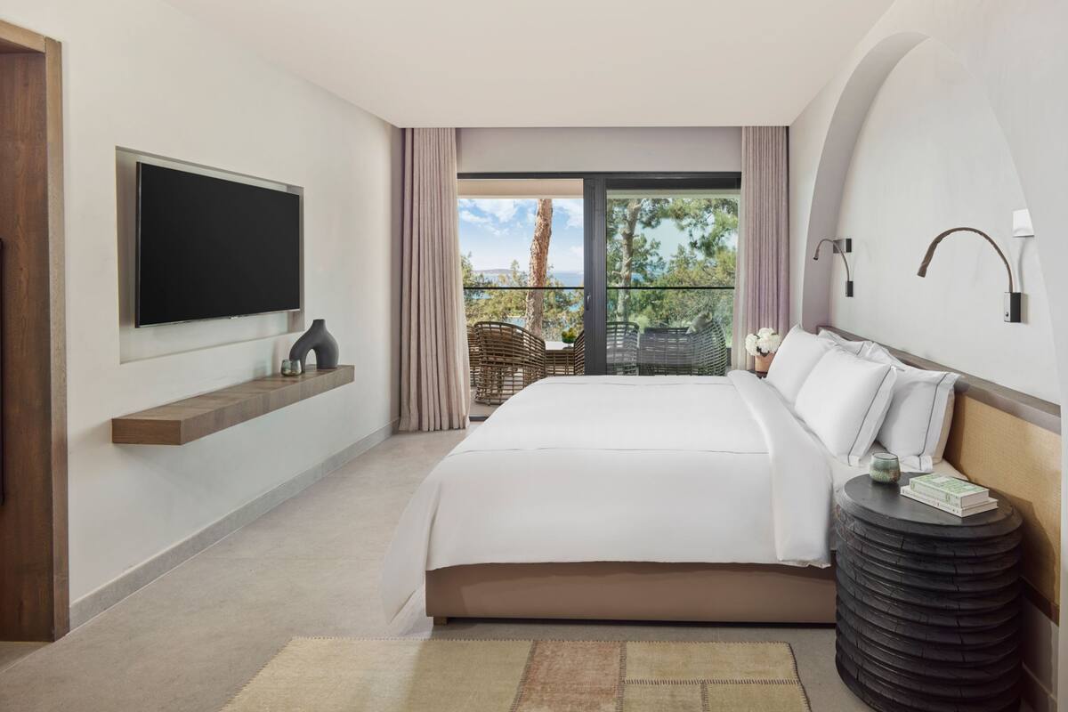 A modern hotel room with a large bed, a flat-screen TV, a small table, and a view of the outdoors.