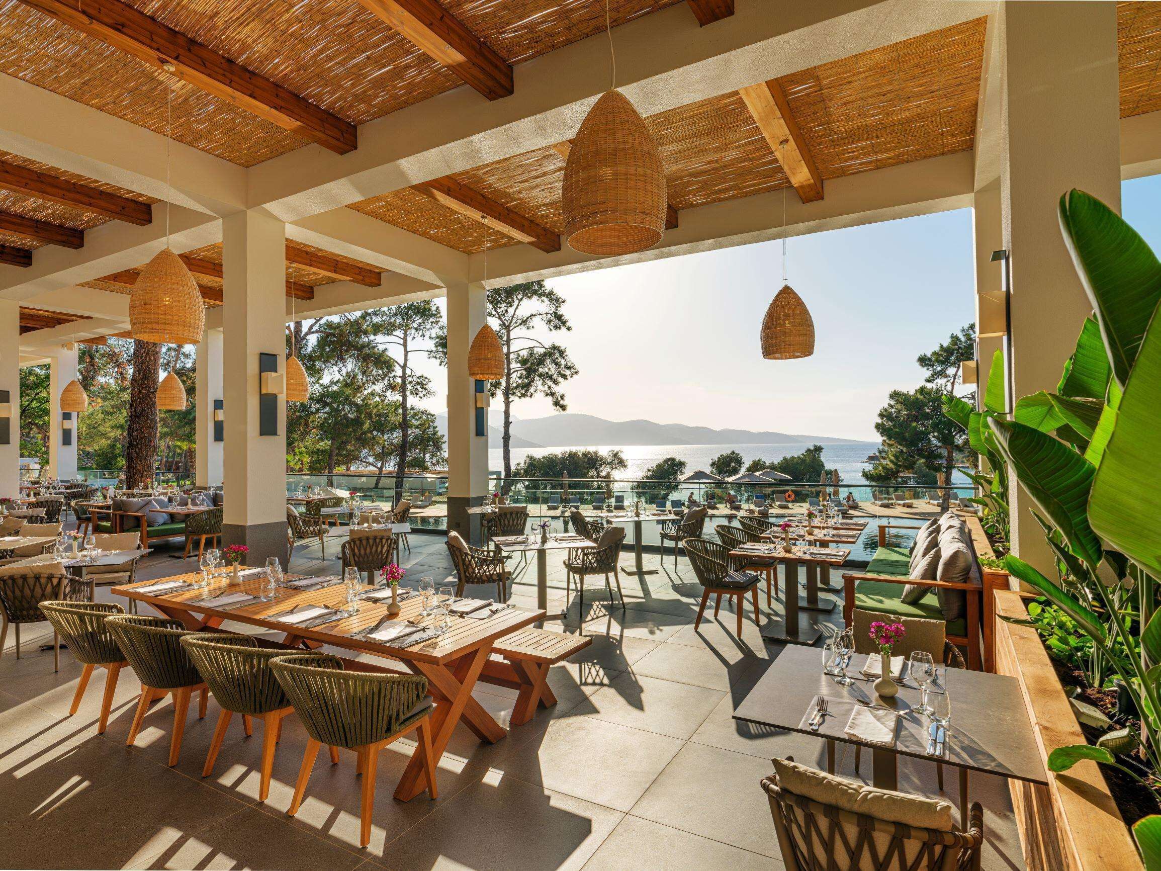 An outdoor dining area with wooden tables and chairs, hanging wicker lamps, and a view of the sea.