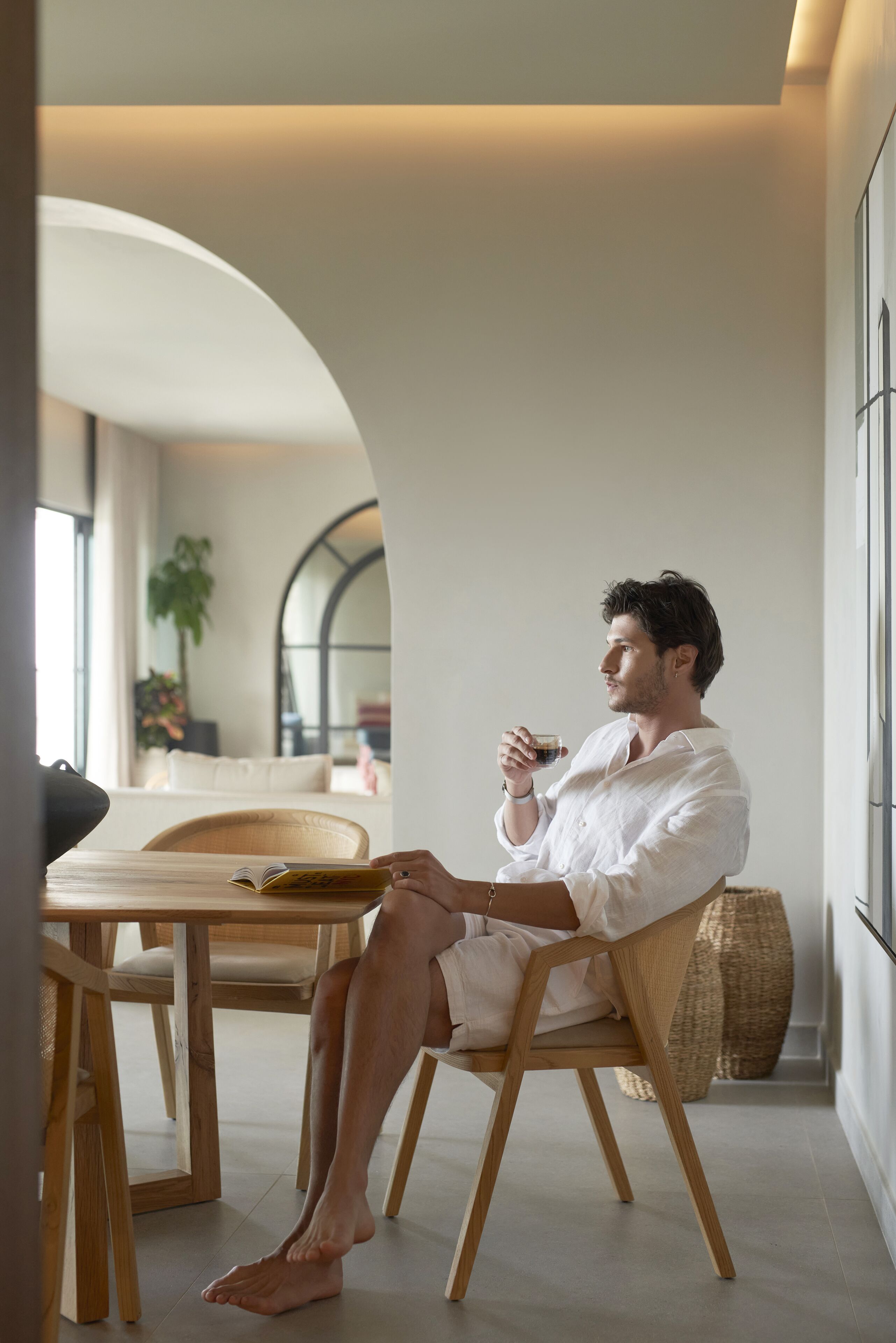 A man sitting at a wooden table in a modern interior with a large window, a plant, and a woven basket.