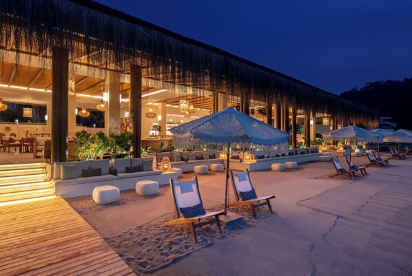 Un complexe hôtelier en bord de mer doté d’une structure à toit de chaume, d’un espace extérieur aménagé et de chaises longues.