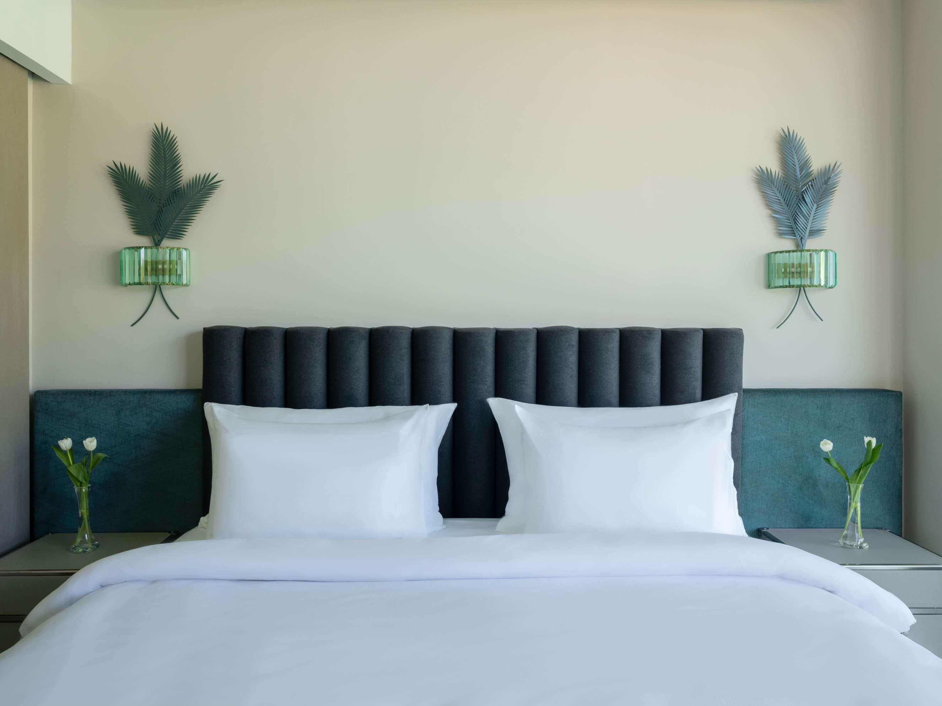 Une chambre d’hôtel avec un grand lit, deux tables de chevet, une tête de lit rembourrée de couleur sombre et des appliques murales décoratives.