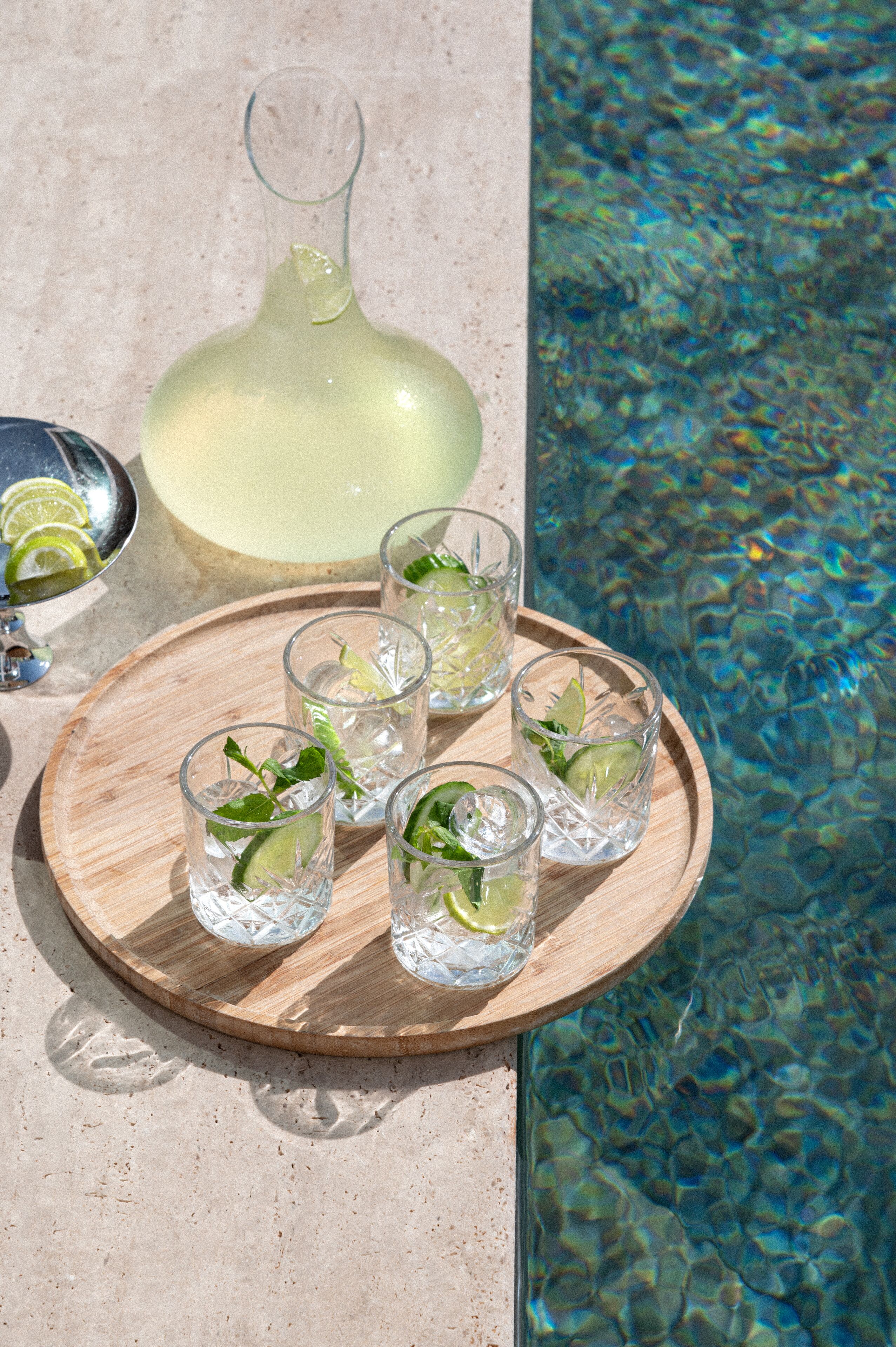 Una bandeja con vasos de una bebida transparente, rodajas de lima y una jarrón, ubicada junto a una piscina con agua cristalina.