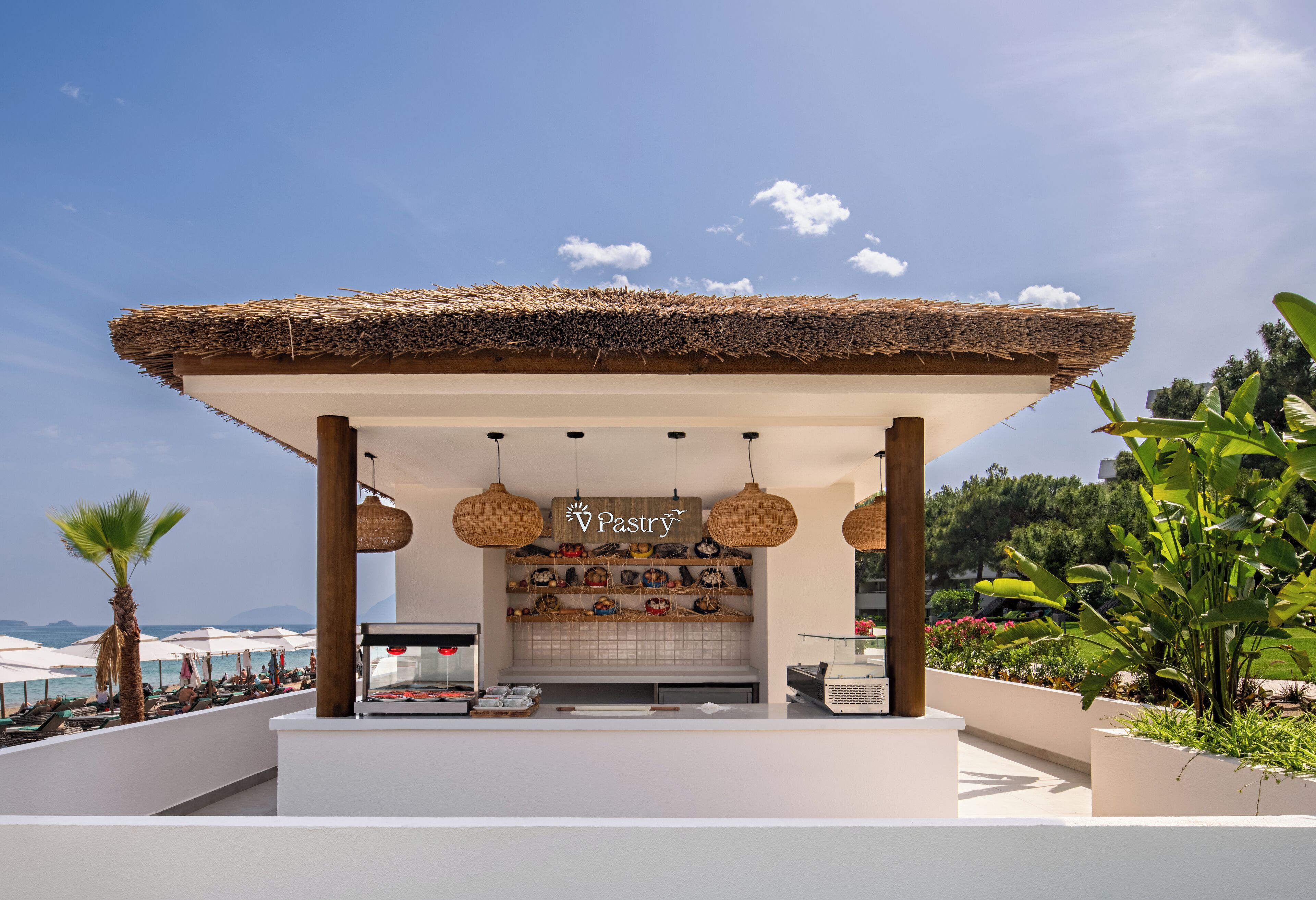 A beachside kiosk with a thatched roof, a sign reading 'Pastry', and a view of the sea.