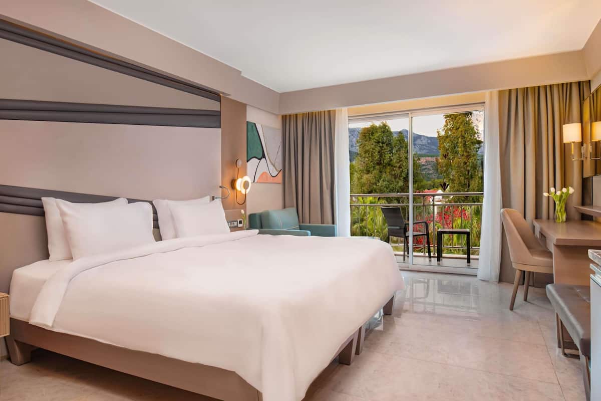 A hotel room with a large bed, a desk, a chair, and a balcony with a view of trees and mountains.