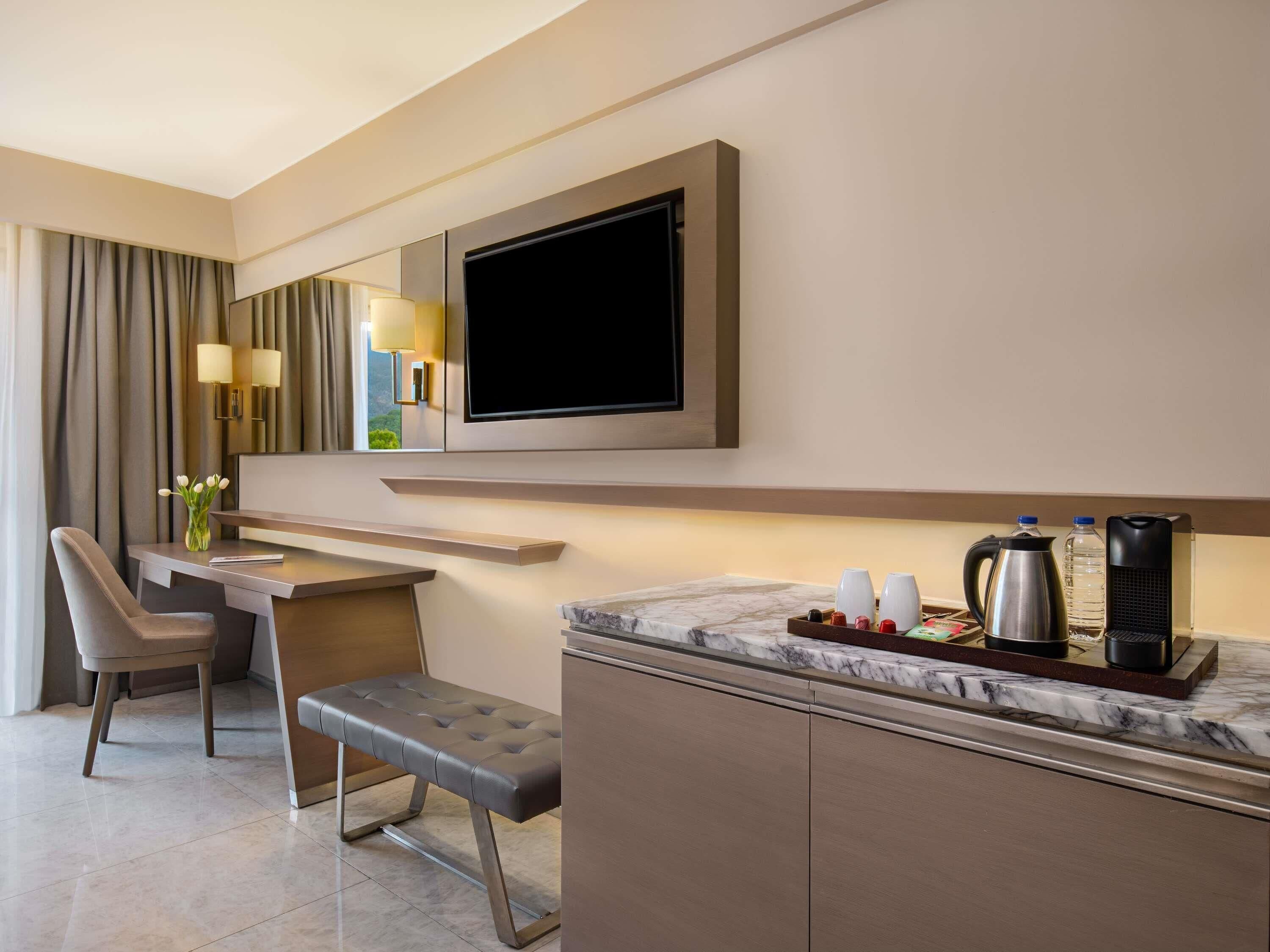 Habitación de hotel moderna con una televisión de pantalla plana, un escritorio con una silla, un banco y una encimera de mármol con cafetera y tazas.