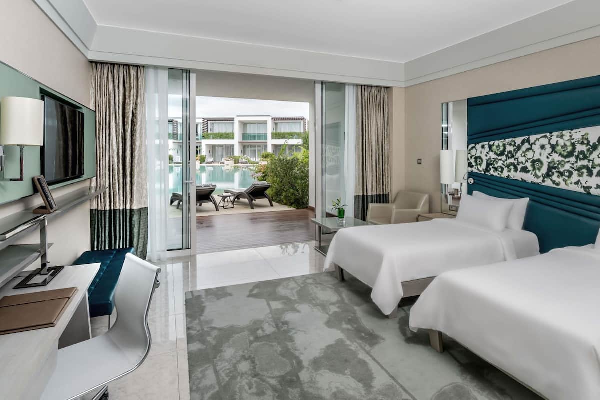 A hotel room with a bed, a desk, a TV, and a view of a pool and building.