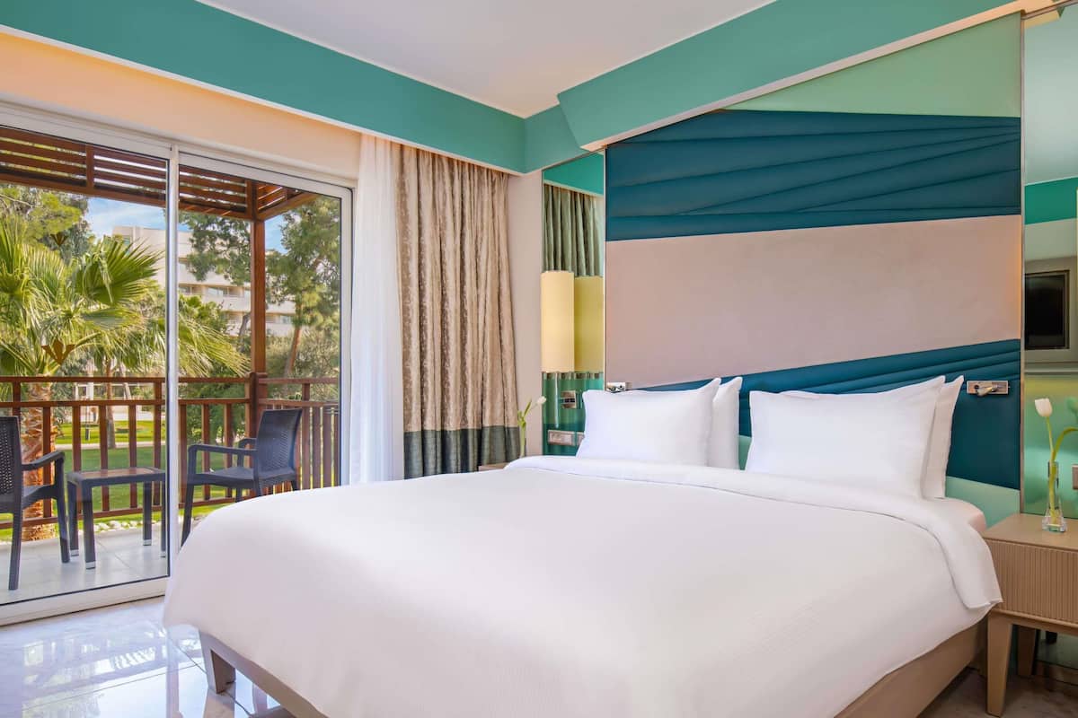 A hotel room with a large bed, a balcony with chairs, and a view of palm trees.
