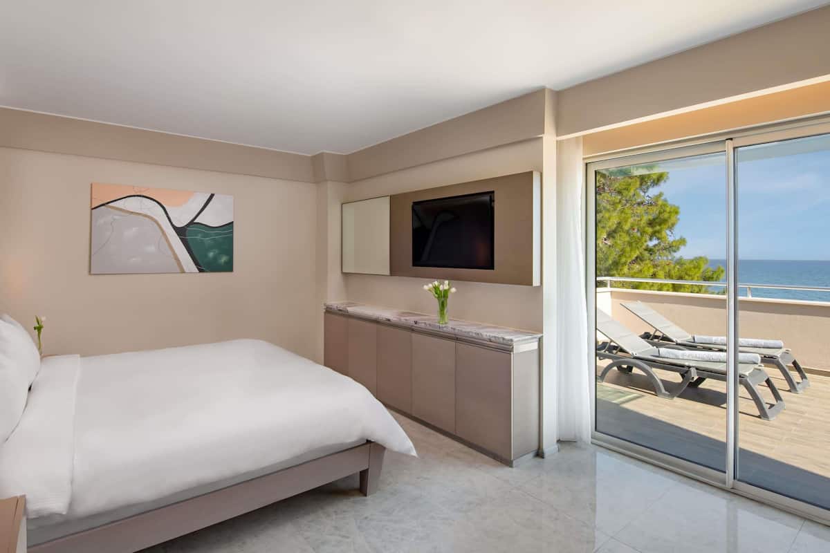 A modern hotel room with a bed, a TV, a bathroom vanity, and a balcony with a view of the sea.