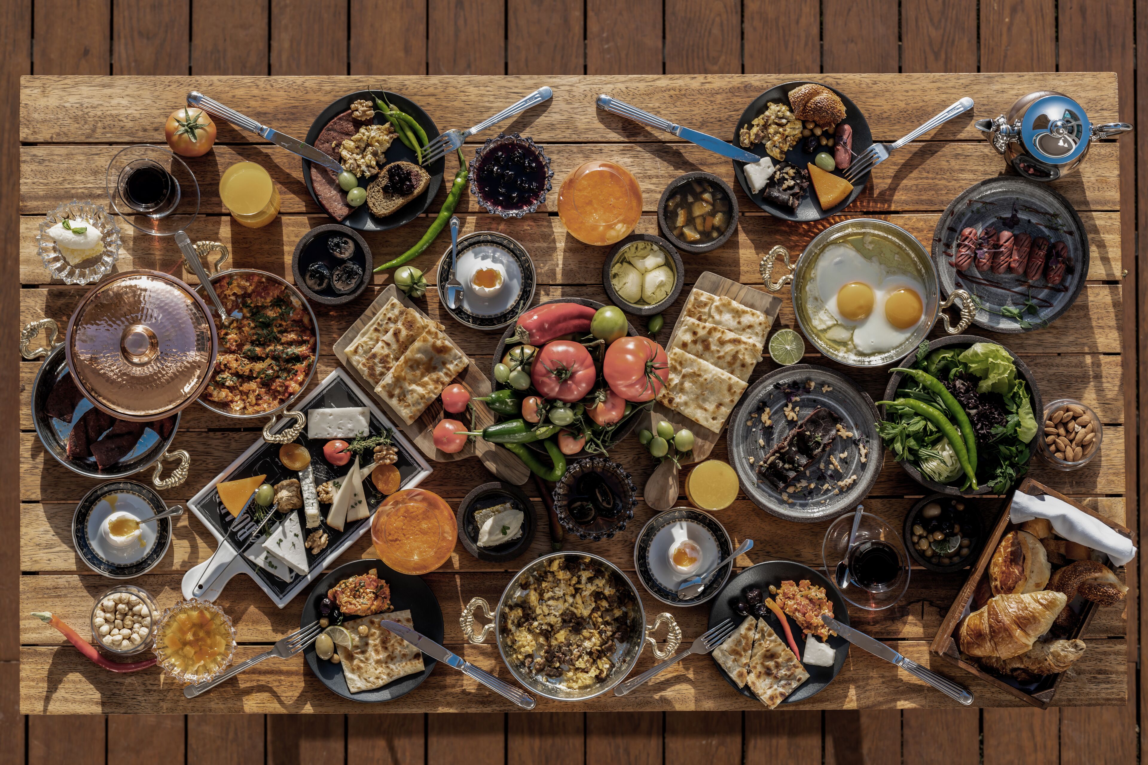 Une table en bois, sur laquelle on trouve une variété d’aliments et de boissons, notamment du pain, des œufs, des tomates et des olives.