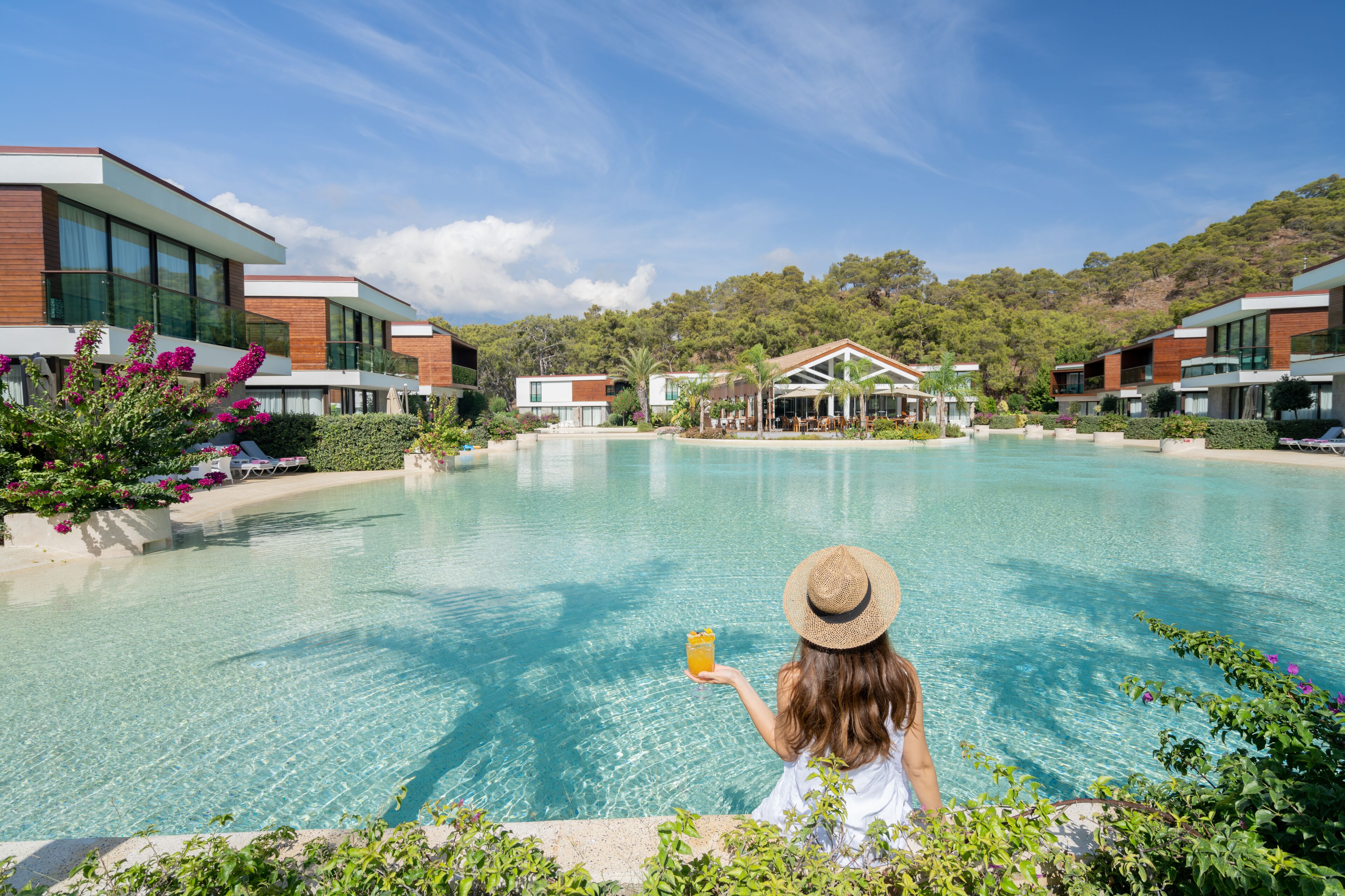 A modern hotel with a pool, surrounded by greenery and a clear sky.