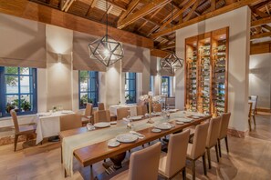 A dining area with a long wooden table set for a meal, surrounded by chairs, with a wine rack and pendant lights above.
