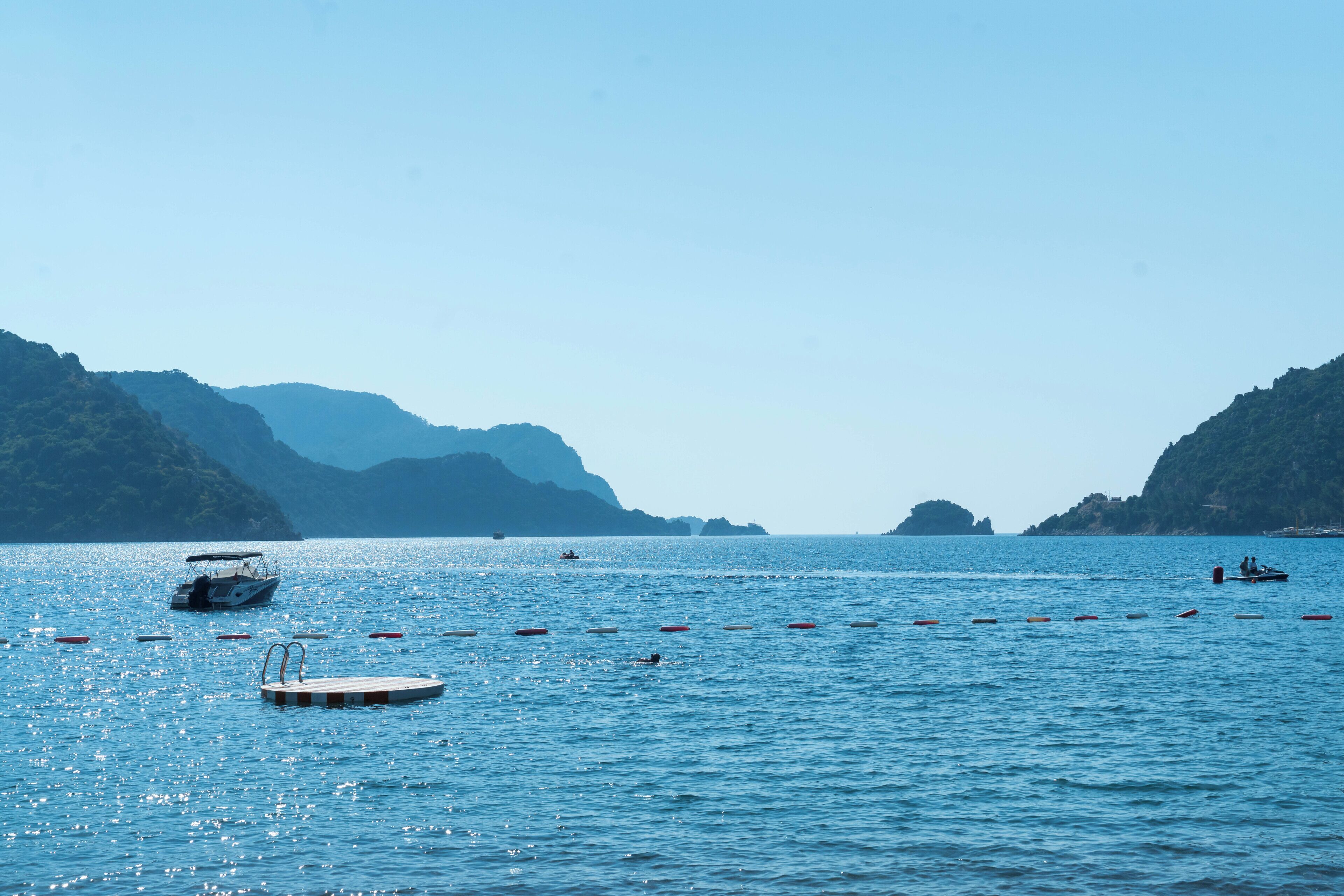 A calm sea with boats and buoys, surrounded by hills.