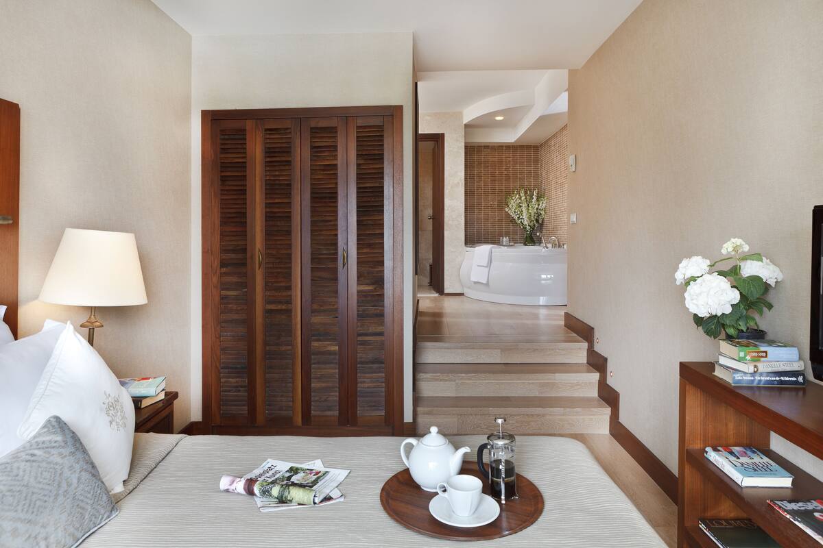 Quarto de hotel com cama, mesa de cabeceira com abajur, bandeja com chaleira, chávenas e um livro. Há uma porta de madeira e uma escadaria que leva a uma casa de banho com lavatório e uma planta.