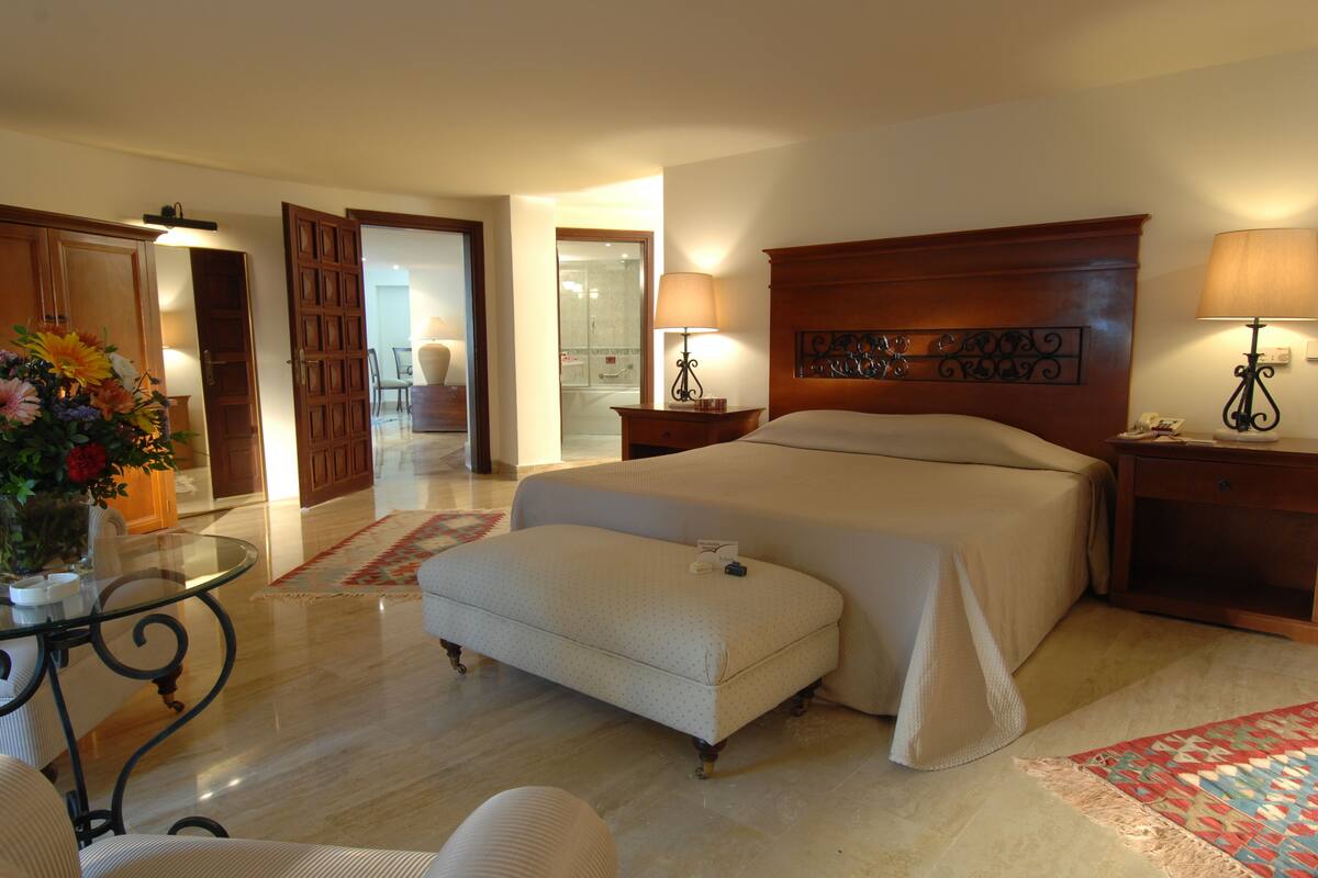 Quarto de hotel com uma cama grande, área de estar com uma mesa e cadeiras, cabeceira de madeira e mesa de cabeceira com abajur.