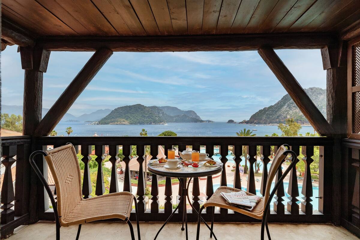 Varanda com cadeiras de vime, mesa com itens de café da manhã e vista para o mar e montanhas.