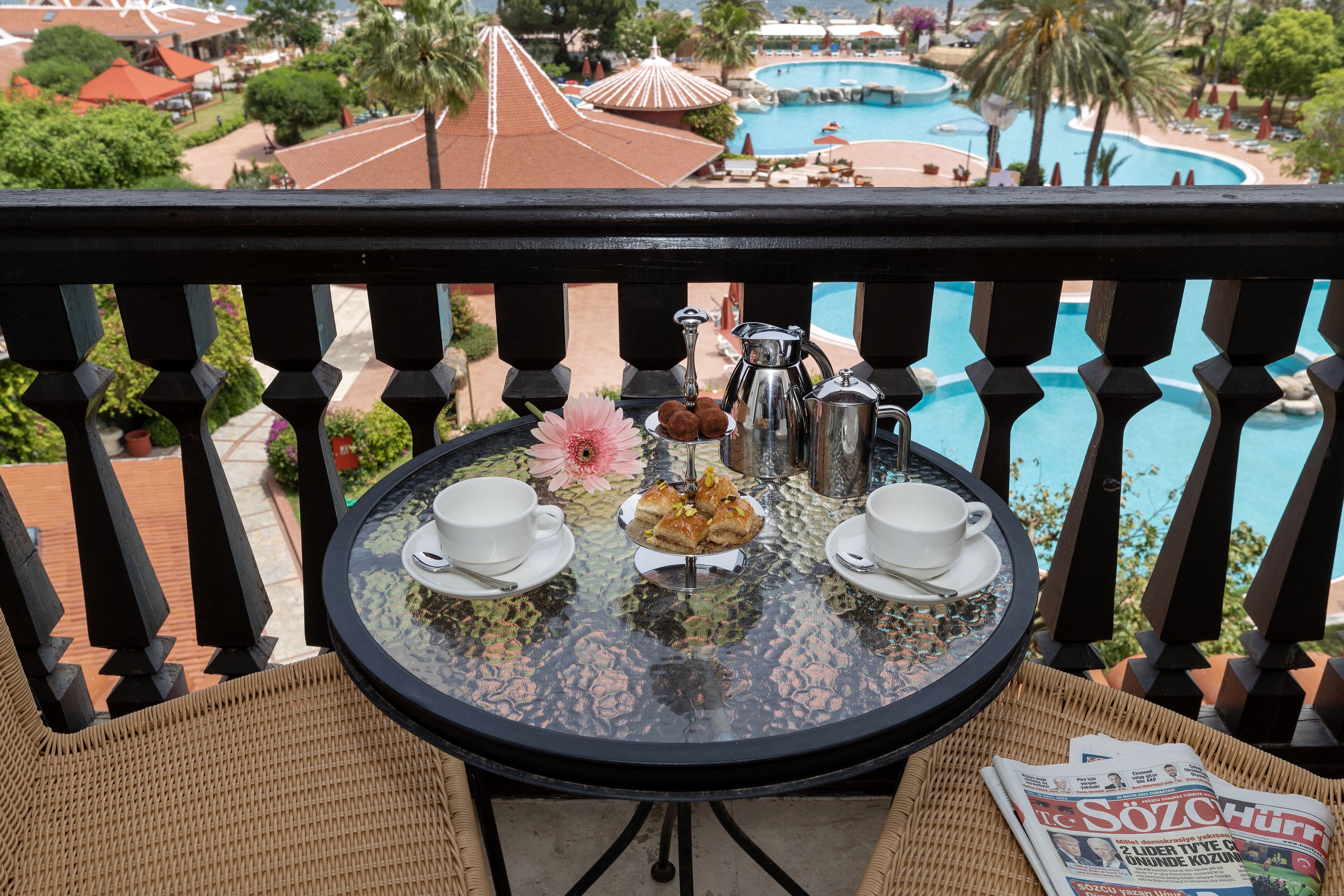 A balcony with a round table, two white cups, a tray of pastries, a silver pot, and a newspaper.