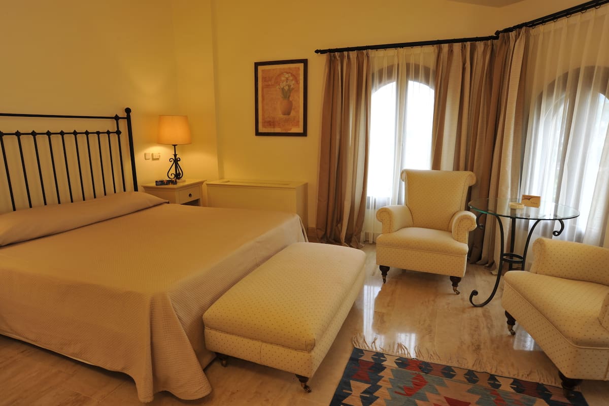 Quarto de hotel com uma cama grande, duas poltronas, uma mesinha e um abajur.