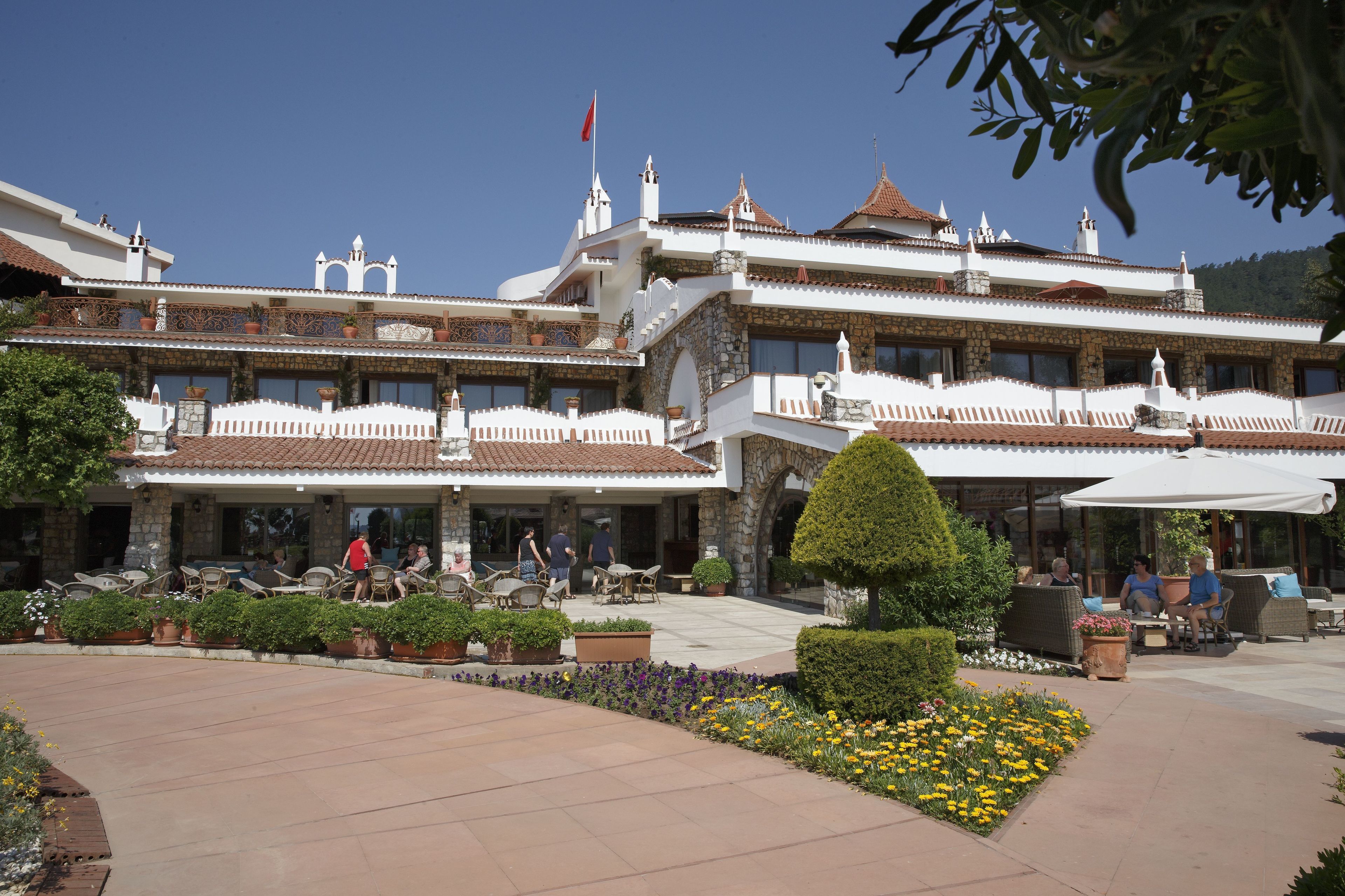 Un edificio grande de varios pisos con estilo arquitectónico tradicional, que cuenta con balcones, una bandera y un jardín bien cuidado con flores y arbustos.