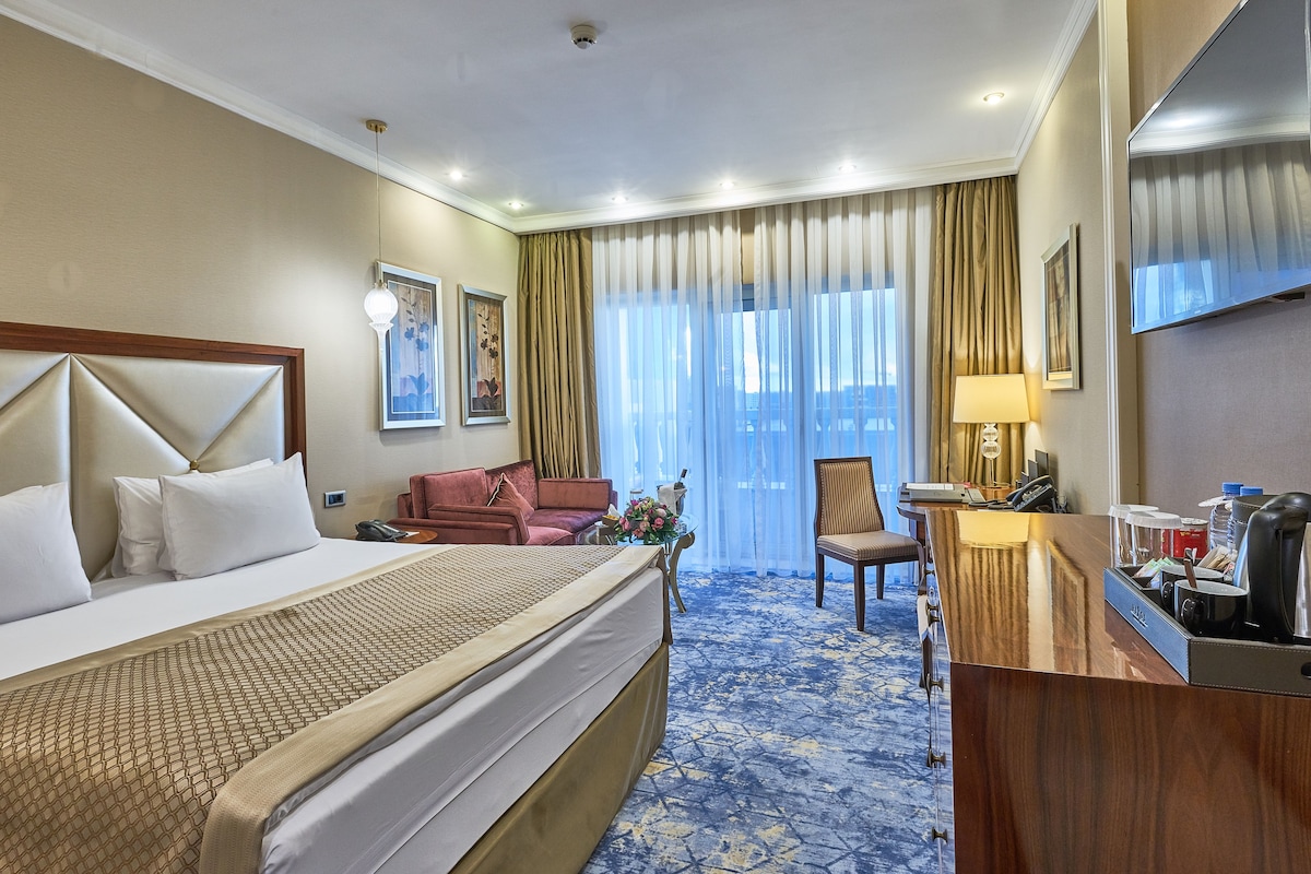A hotel room with a large bed, a desk with a lamp, a chair, and a view of the city through large windows.