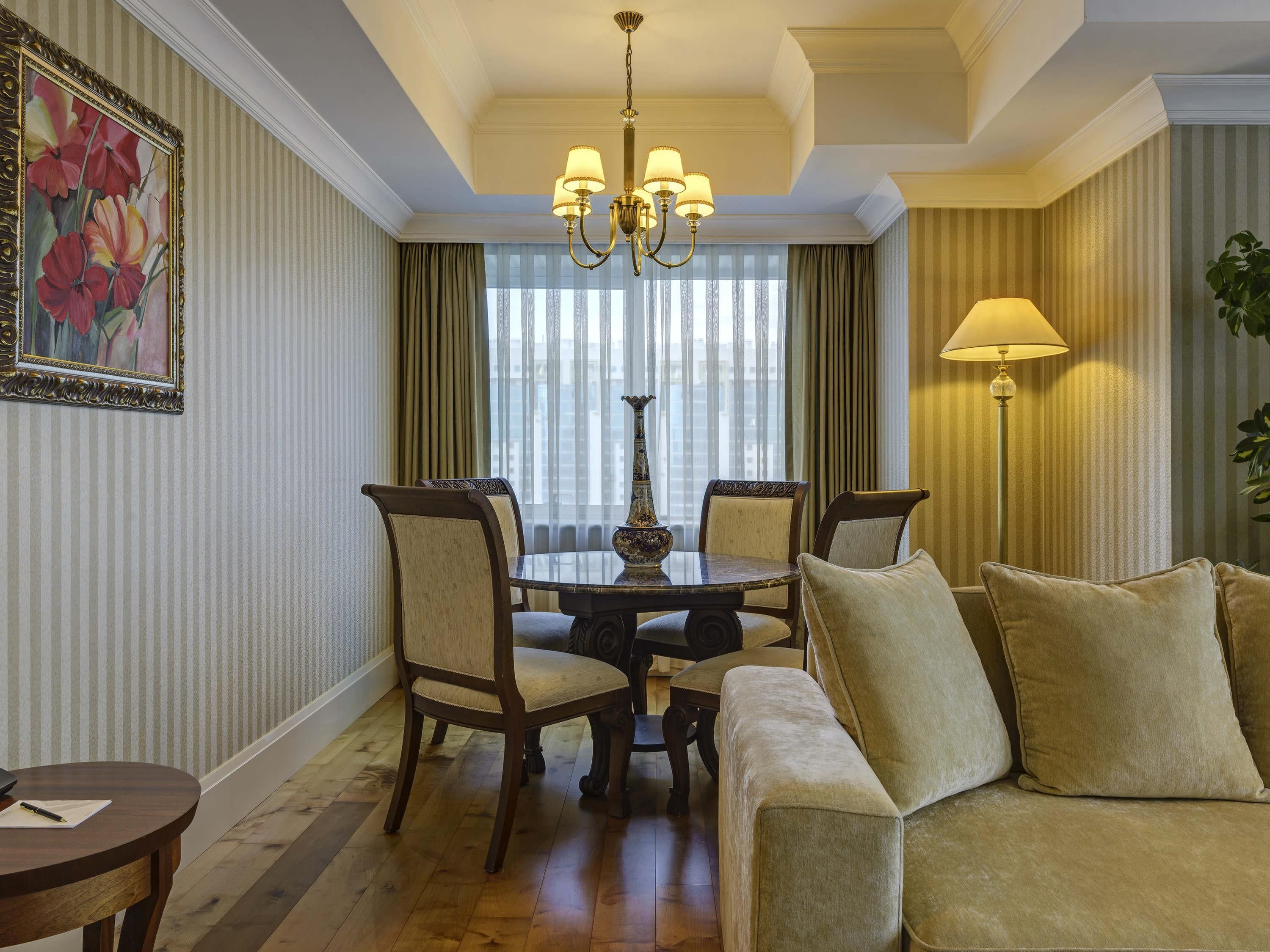 Habitación de hotel con una mesa de comedor, sillas, un sofá, una araña y un amplio ventanal con cortinas.