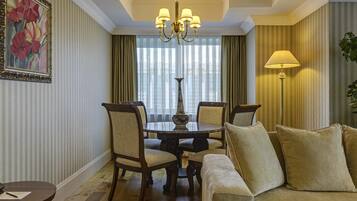 Habitación de hotel con una mesa de comedor, sillas, un sofá, una araña y un amplio ventanal con cortinas.