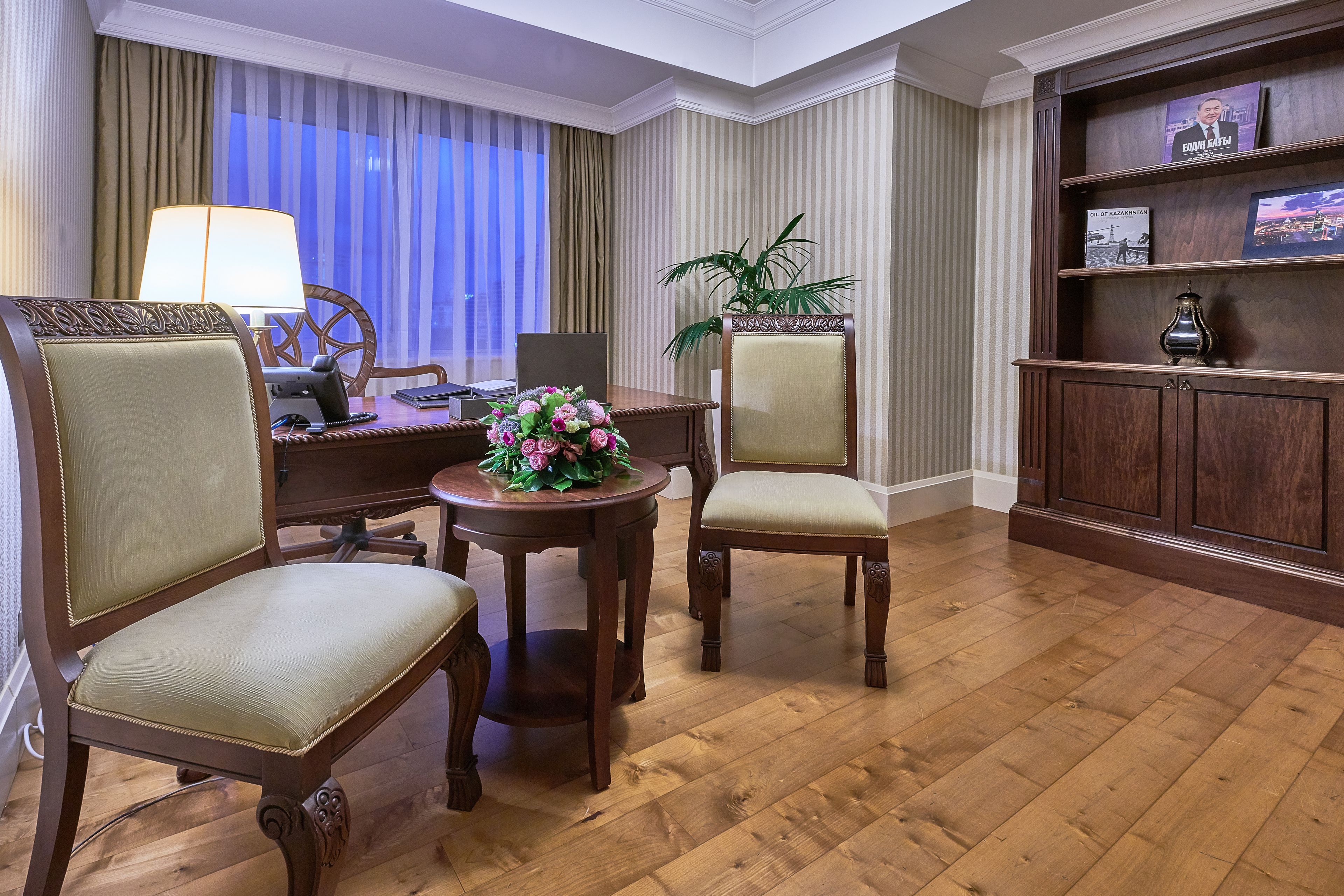 Quarto de hotel com piso de madeira, uma escrivaninha com abajur, duas cadeiras, uma pequena mesa com flores, uma estante de livros e uma grande janela com cortinas.