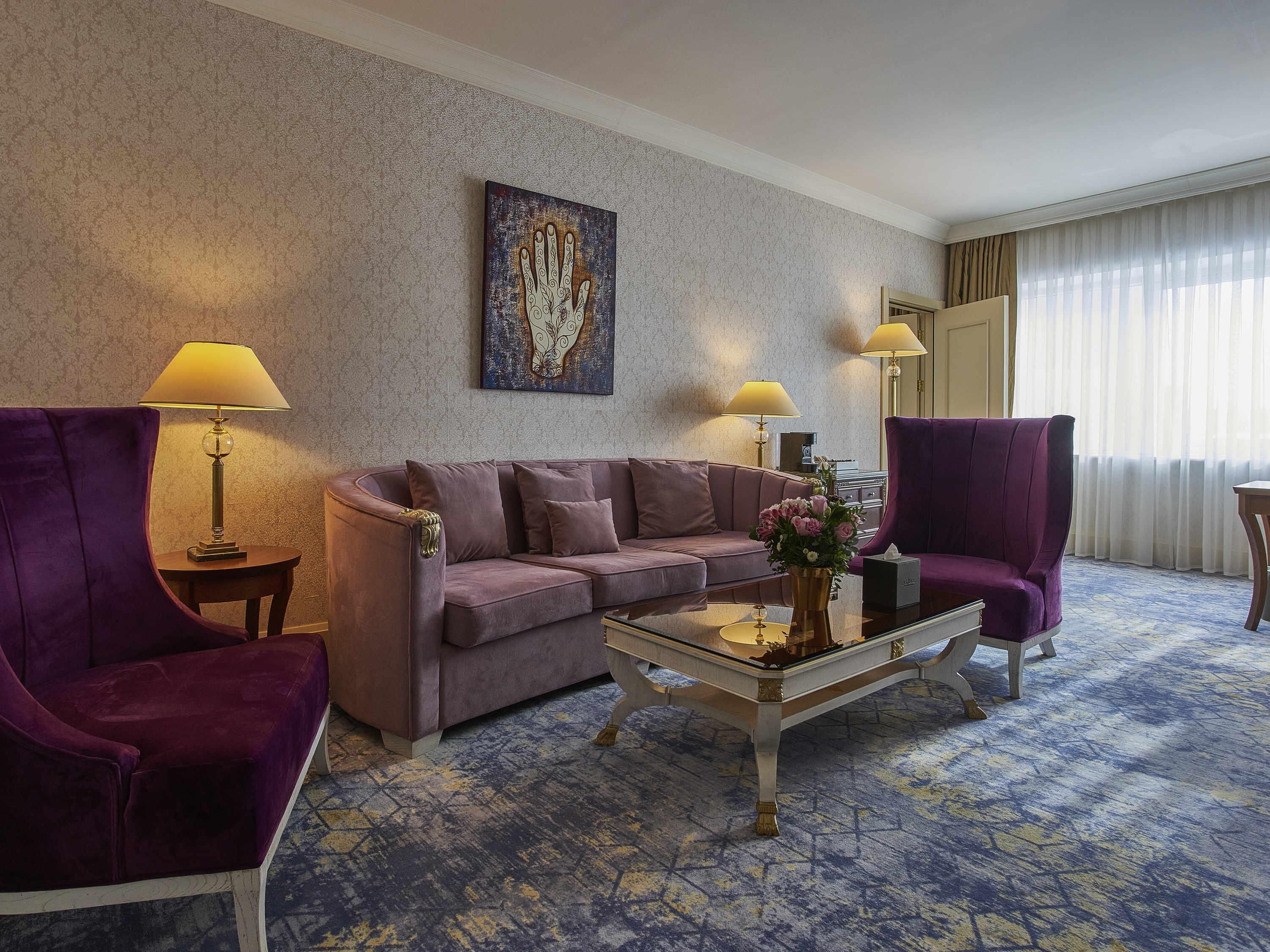 Habitación de hotel con un sofá púrpura, dos sillones, una mesita de centro y un cuadro en la pared.