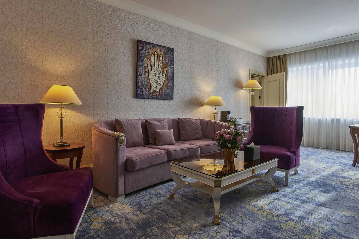 A hotel room with a purple sofa, two armchairs, a coffee table, and a painting on the wall.