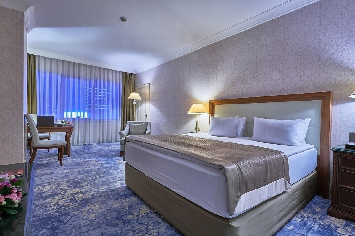 A hotel room with a large bed, a desk, a chair, and a view of the city through the window.