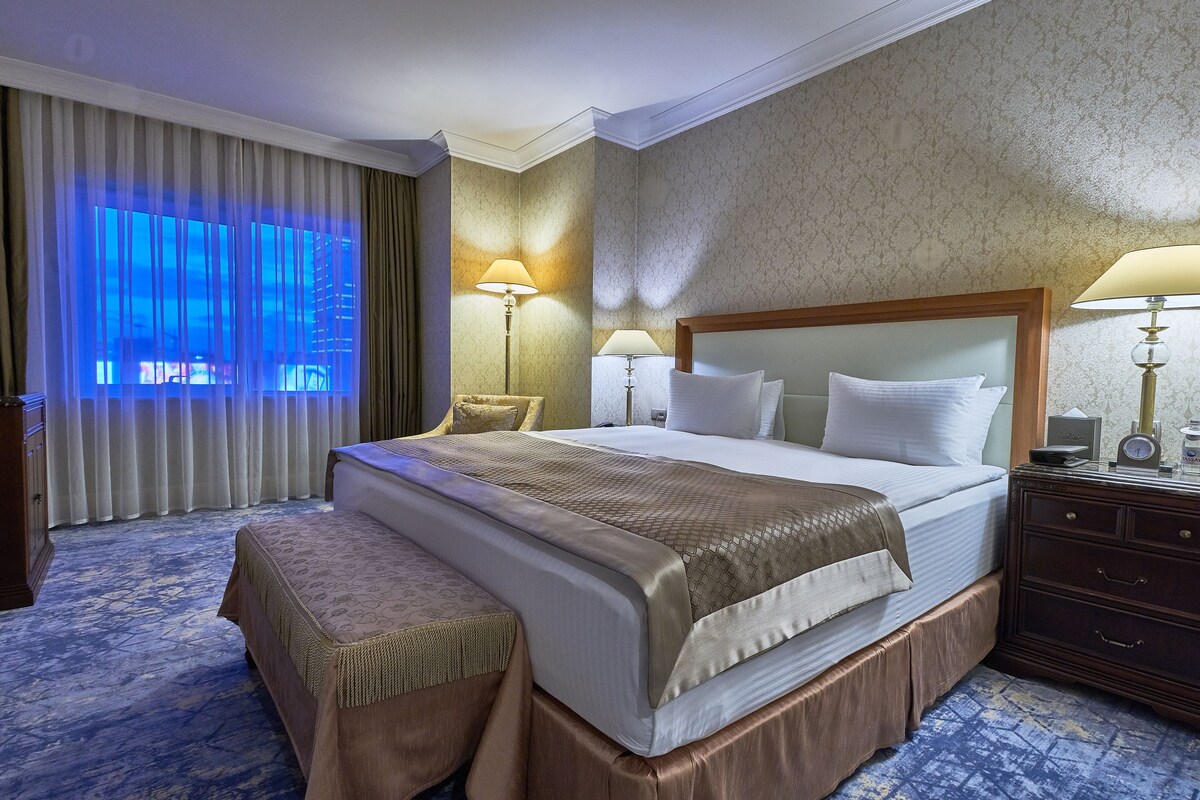 A hotel room with a large bed, two bedside lamps, a wooden nightstand, a bench, and a view of the city through the window.