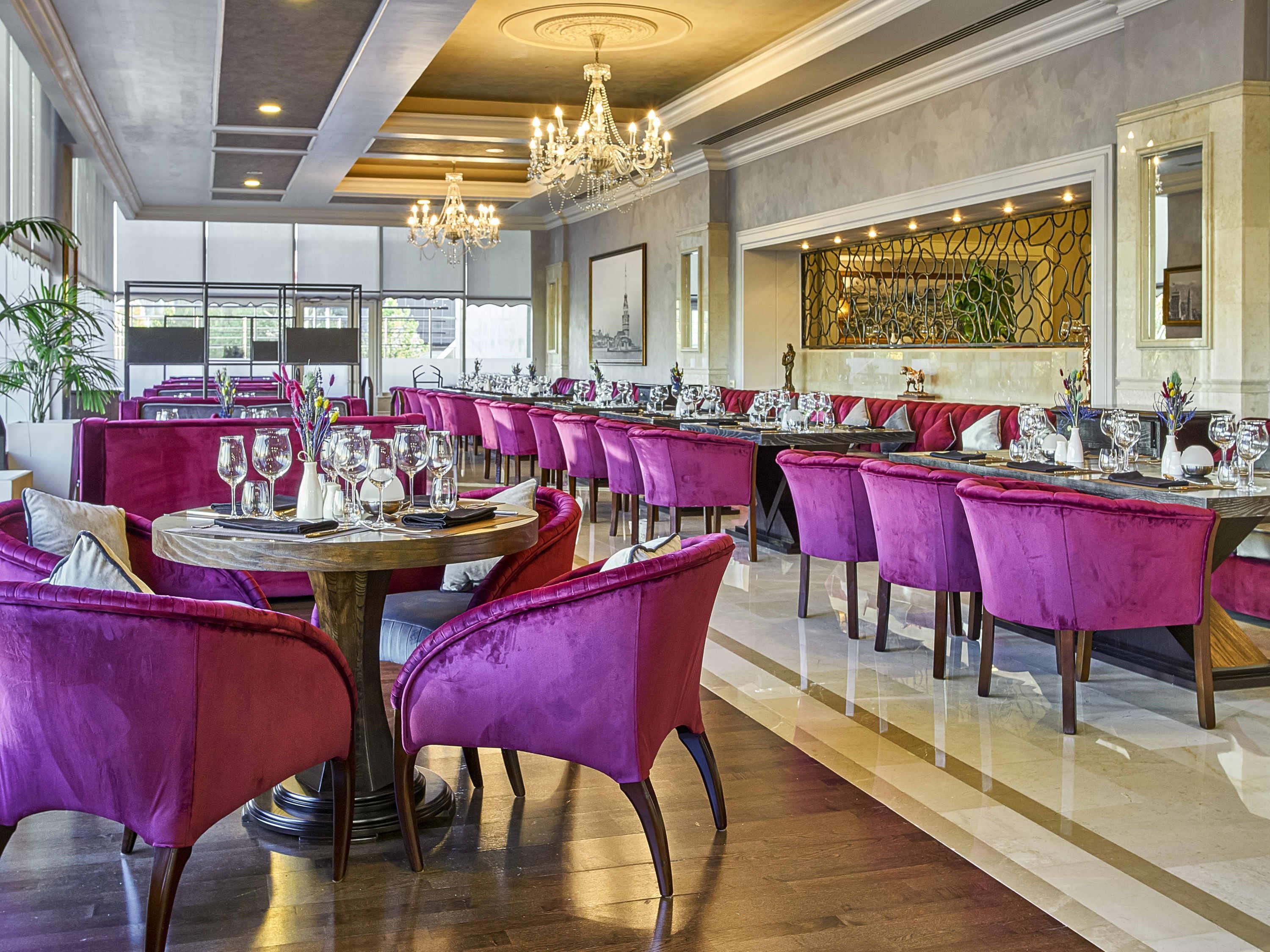 A spacious dining area with round tables, purple upholstered chairs, chandeliers, and a large decorative wall.