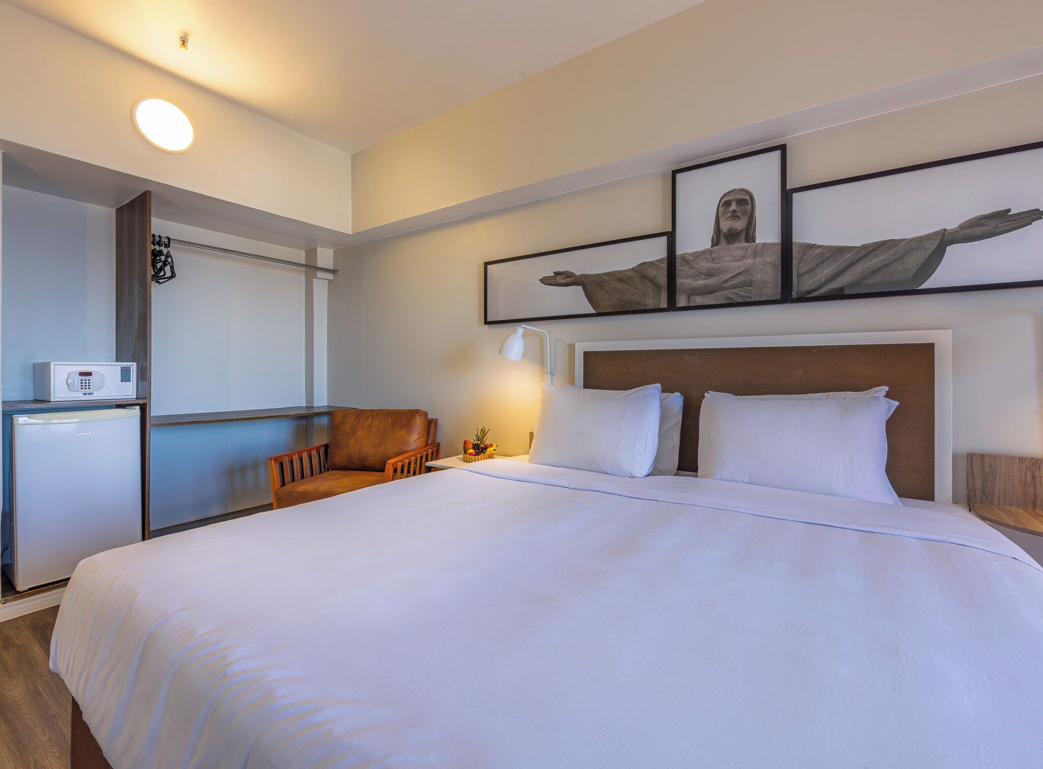 Ett hotellrum med en säng, en stol, ett skrivbord, ett kylskåp och en tavla som hänger på väggen.