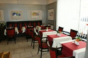 A restaurant with red and white table settings, a central seating area with a black and red bench, and framed artwork on the walls. - Hotel Acon (Düsseldorf)