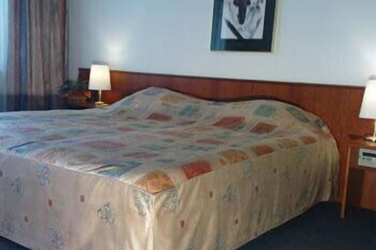 Habitación de hotel con cama, dos lámparas de noche, un cuadro en la pared y una cortina en la ventana.