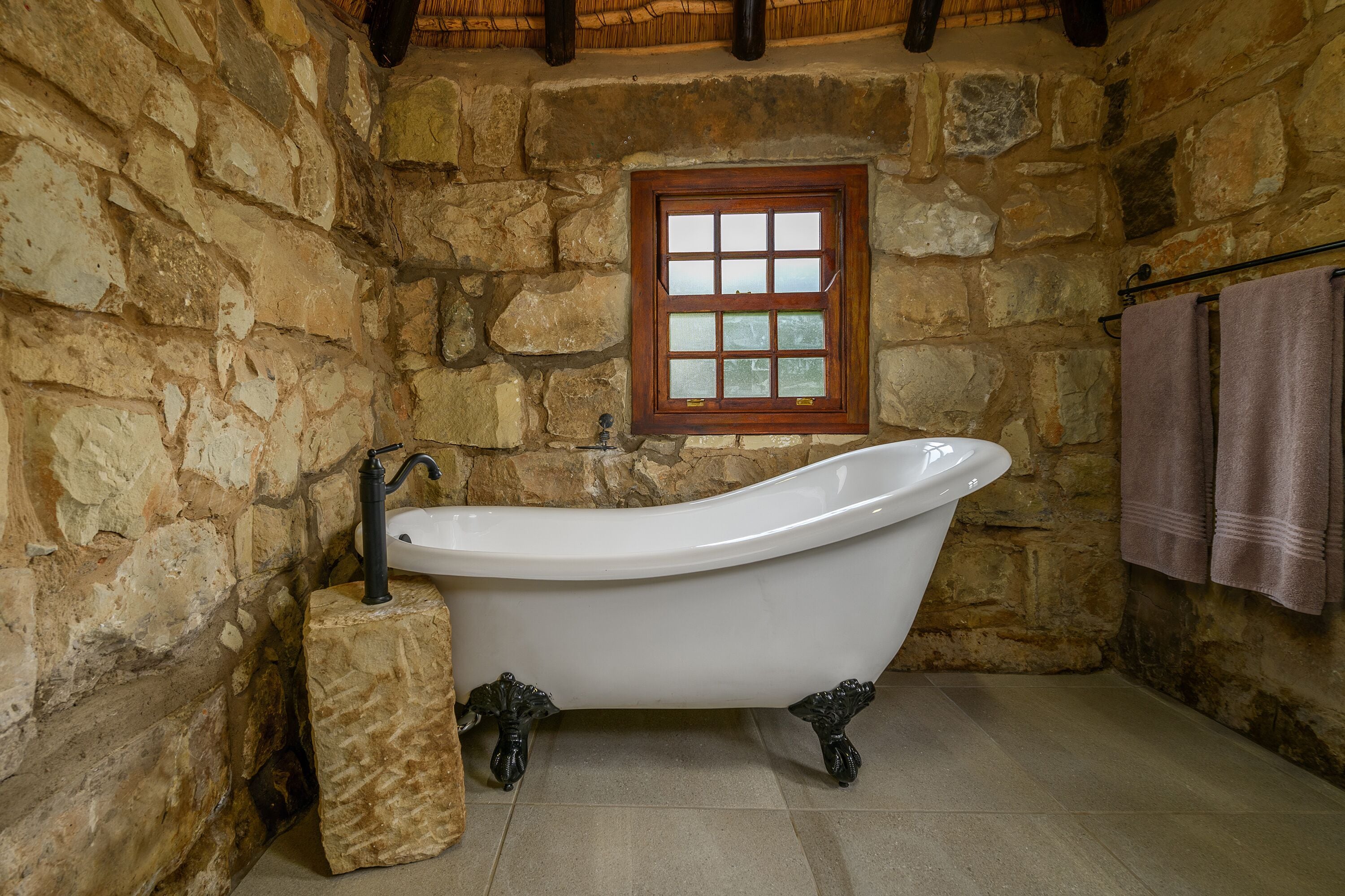 A stone-walled bathroom with a freestanding bathtub, a window, and towels on a rack.